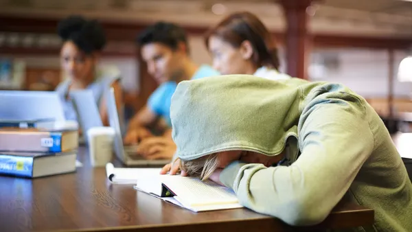 A student sitting at a desk is shown with their head down on a desk and their hood pulled over their head. Other students can be seen working in the background.