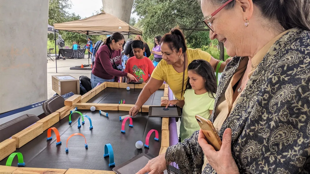 An adult and a child use iPads to control the movement of a ping pong ball through obstacles on table at an outdoor event while another adult moves one of the obstacles.