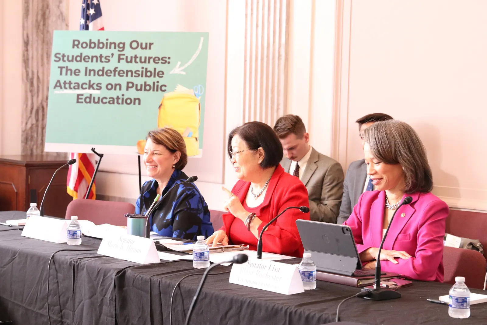 Three people sit side-by-side at a table. There are microphones on the table. A large sign behind and to the right of them says "Robbing our students' futures: The Indefensible attacks on public education"