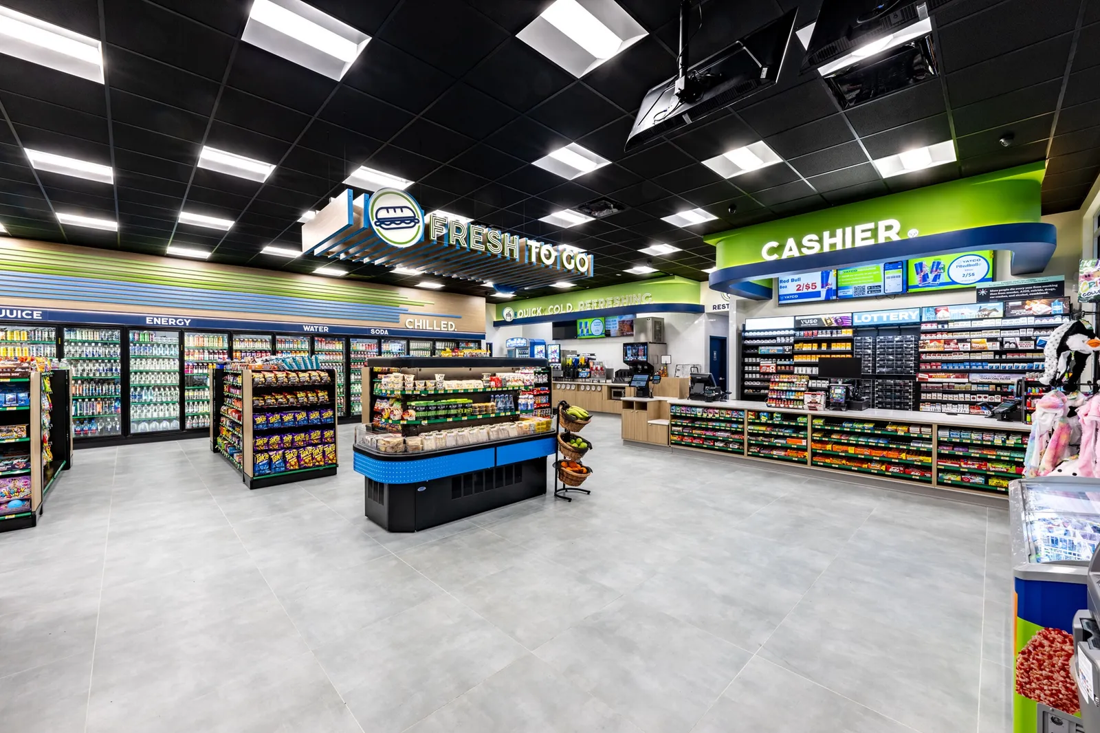 A photo of the interior of a store, showing shelves, cookers and the checkout area. A sign in the center of the store suspended from the ceiling says Fresh To Go, while signs on the walls say Cashier, ice, water, energy, soda, chilled and Chiller. The store is decorated in greens and blues along with wood tones.