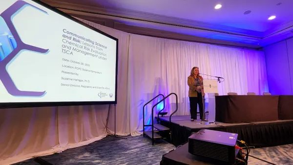 A woman standing behind a podium speaking into a microphone, with a presentation on the left.