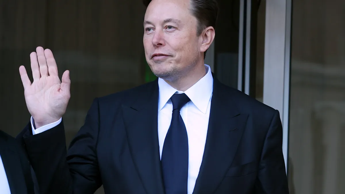 A man in a suit and a navy blue tie waving with his right hand.