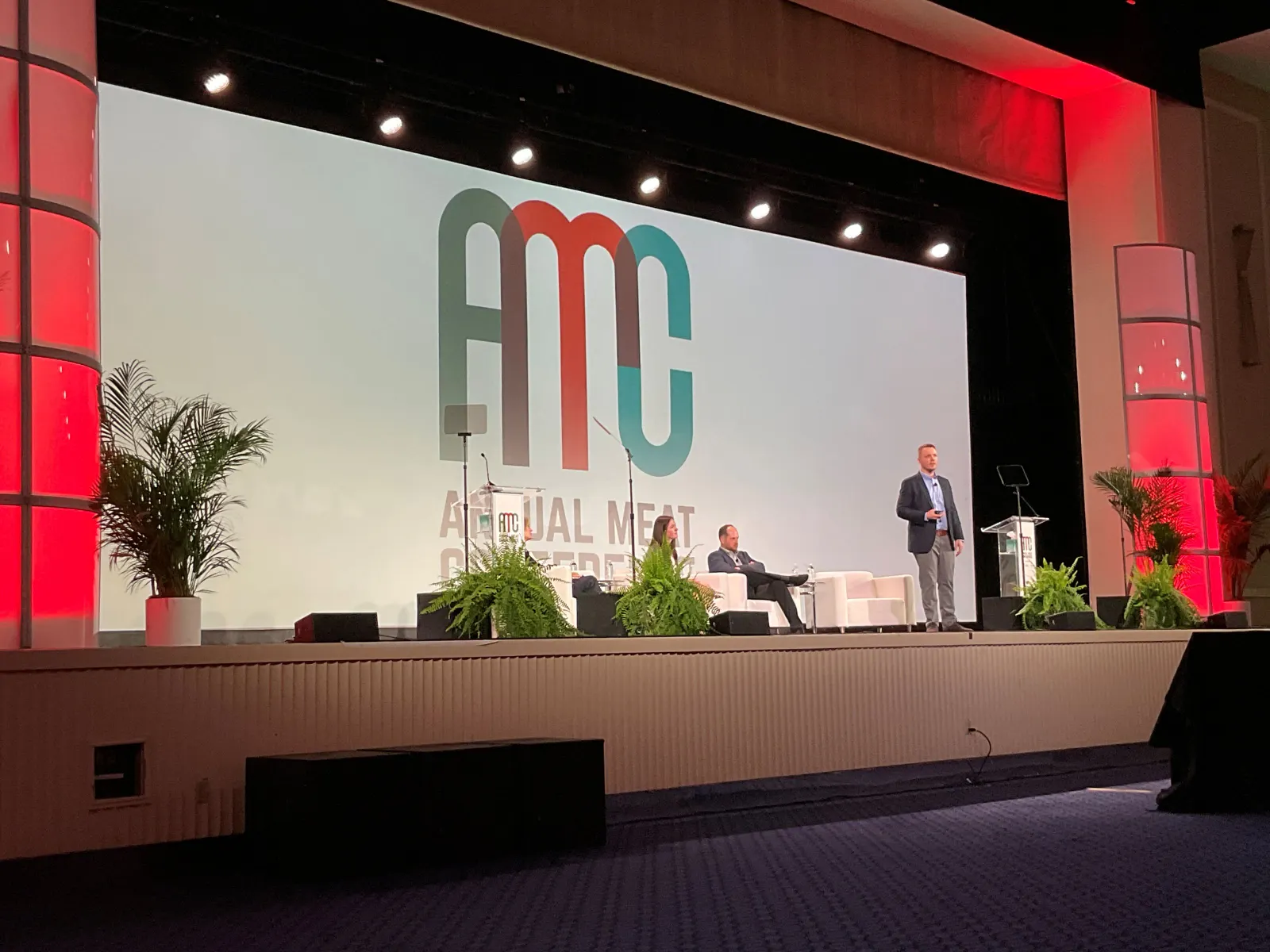 A person speaking on a stage in front of a sign that says "AMC Annual Meat Conference"