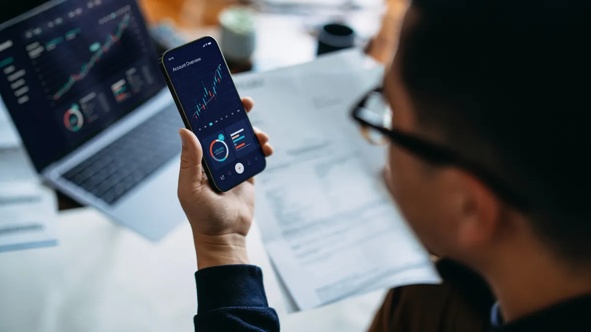 Young Asian man managing finance and investment online, analyzing stock market trades with smartphone and laptop. Making financial plans. Banking and finance, investment, financial trading, mobile banking with technology concept