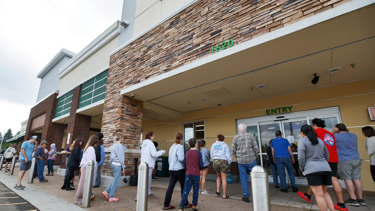 A group of people wait outside a grocery store glass door that has a sign posted on it.