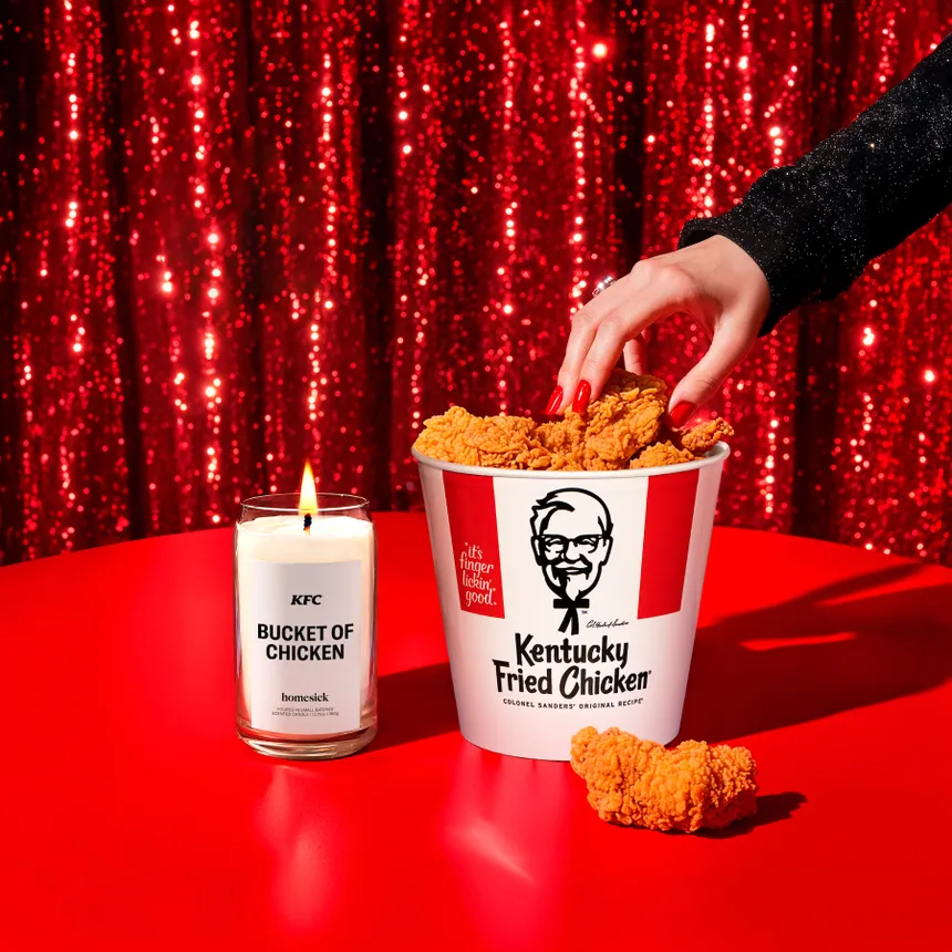 A person reaching into a bucket of KFC fried chicken with a Homesick candle sitting next to it.