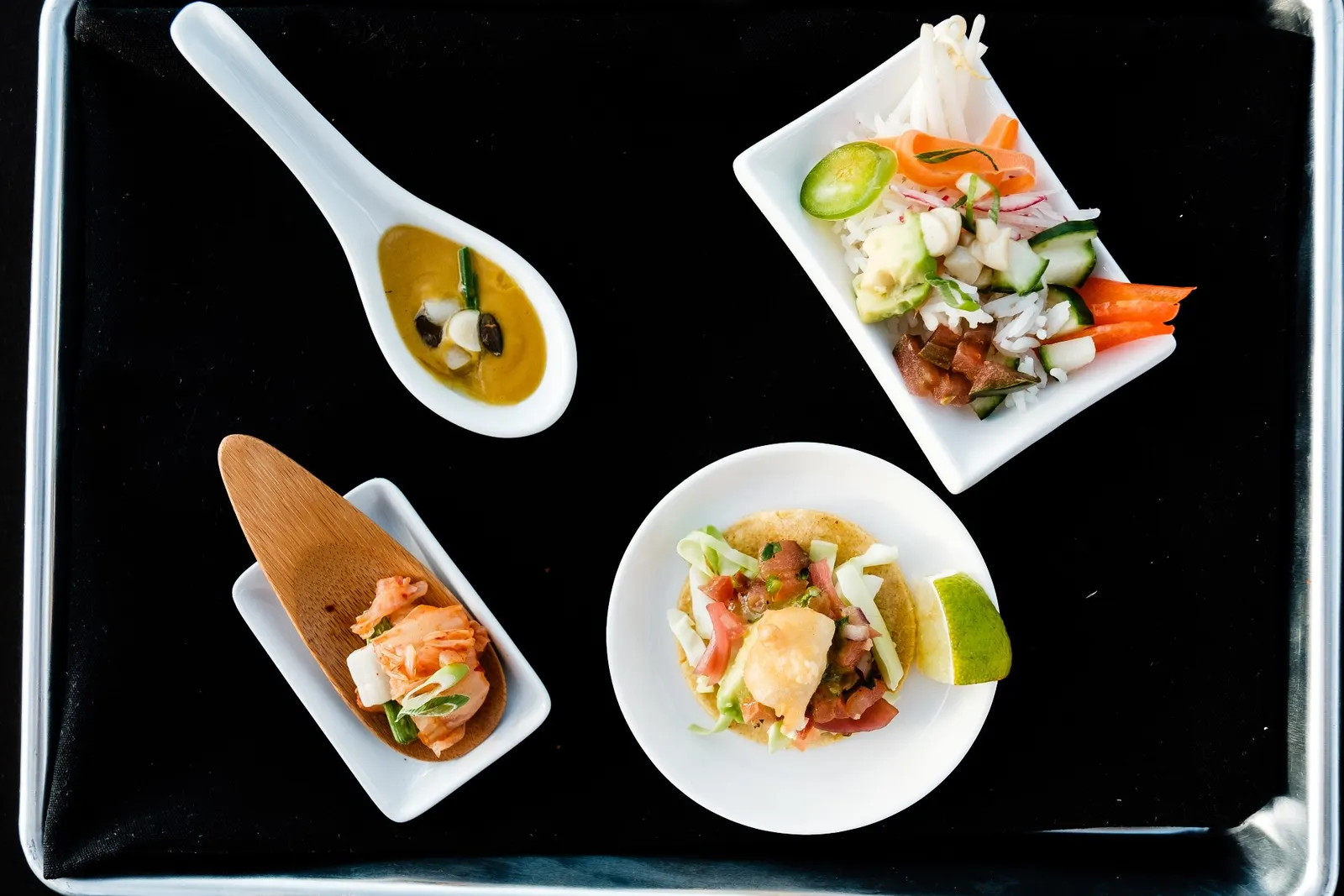 Small white plates of dishes made with cultivated yellowtail, including soup, kimchi, a miniature soft taco and rice on a black tray.