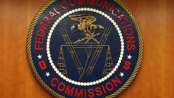 The red, white, blue, and gold seal of the Federal Communications Commission hangs on a wooden wall