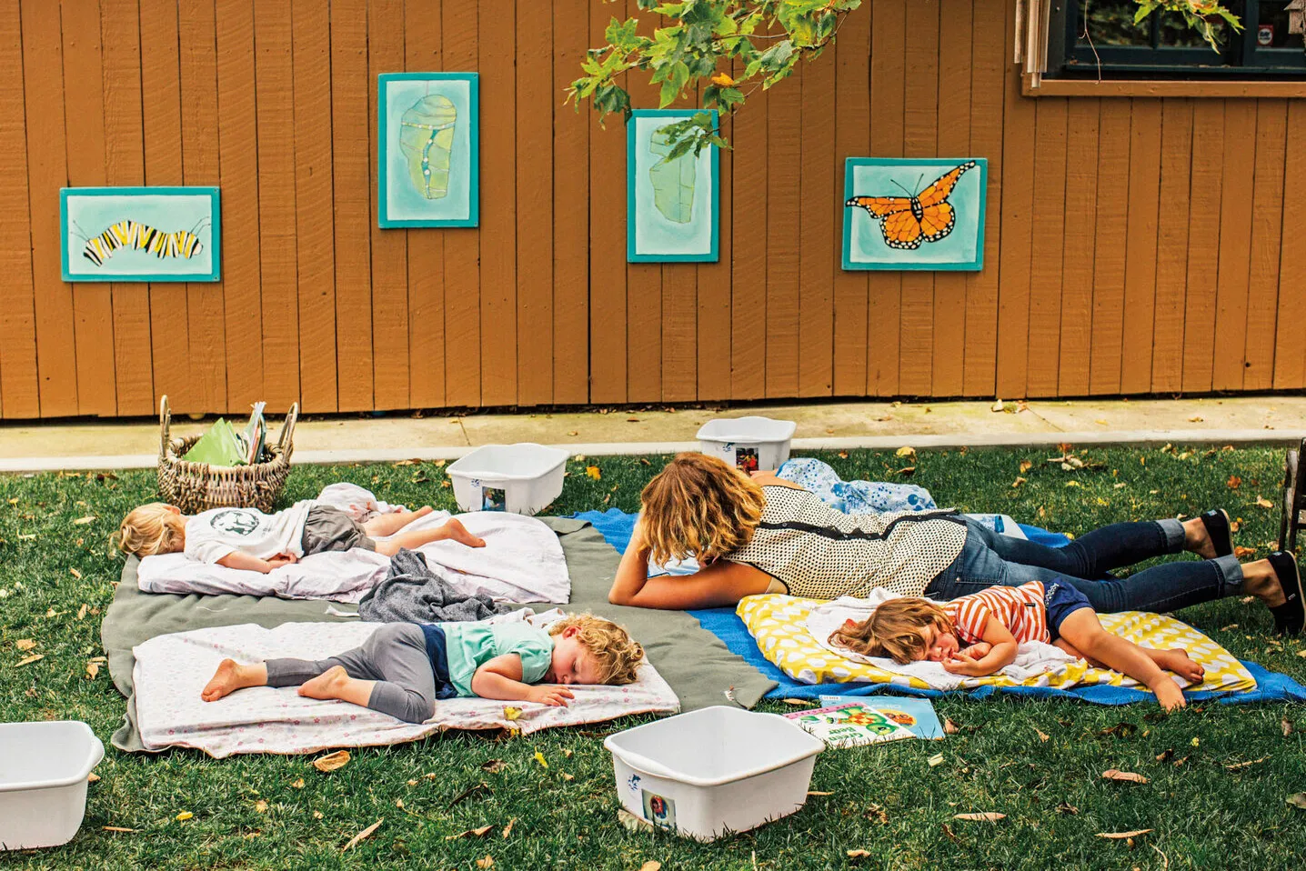 Three children sprawl on blankets in the grass near a caregiver, taking naps.
