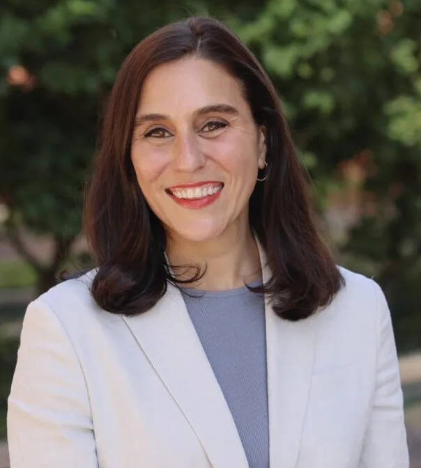 Woman with long dark hair and a white suit smiles at the camera.