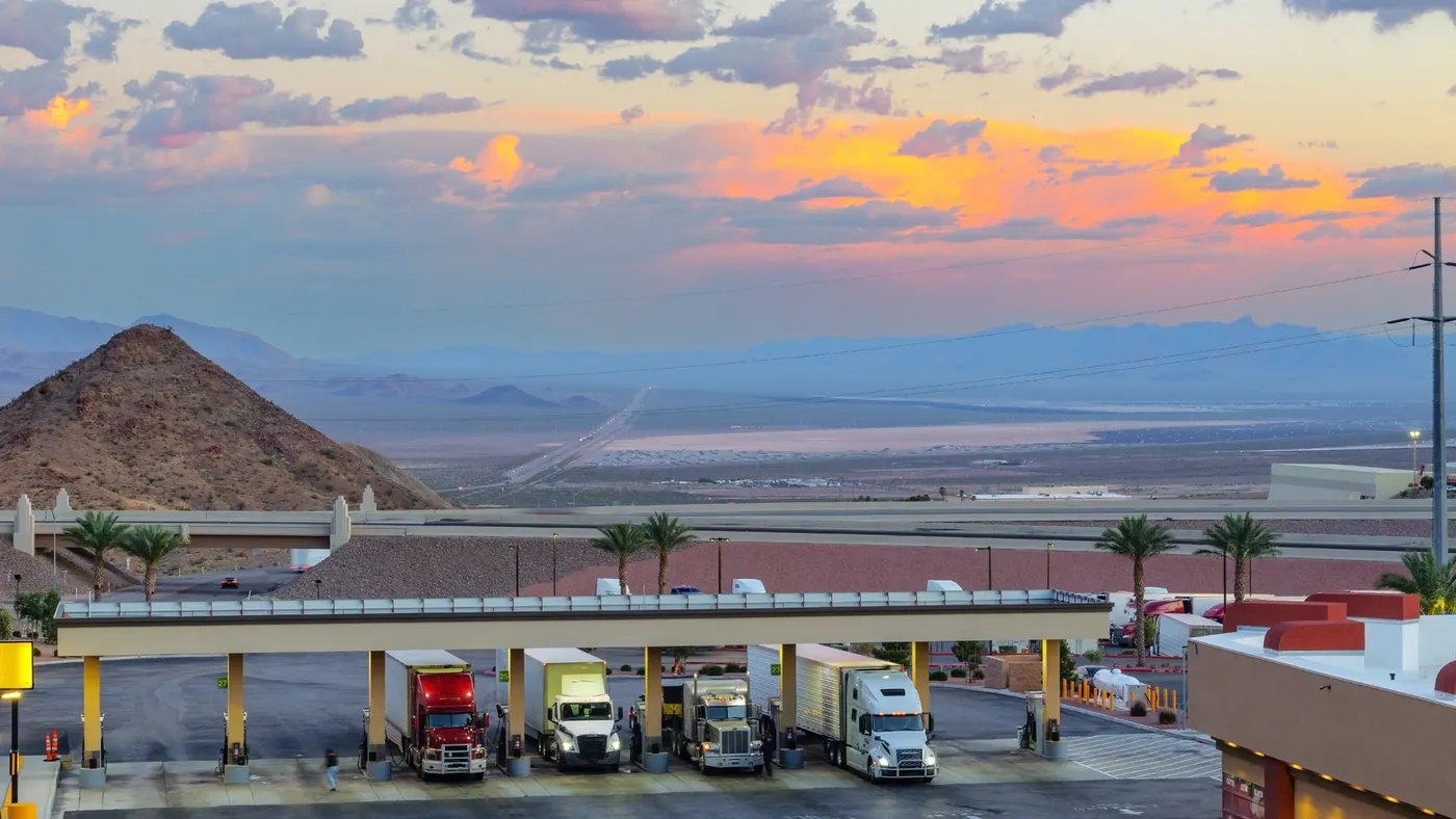 Trucks fueling against a dramatic evening sky showing sunlight across clouds amid a desert landscape.