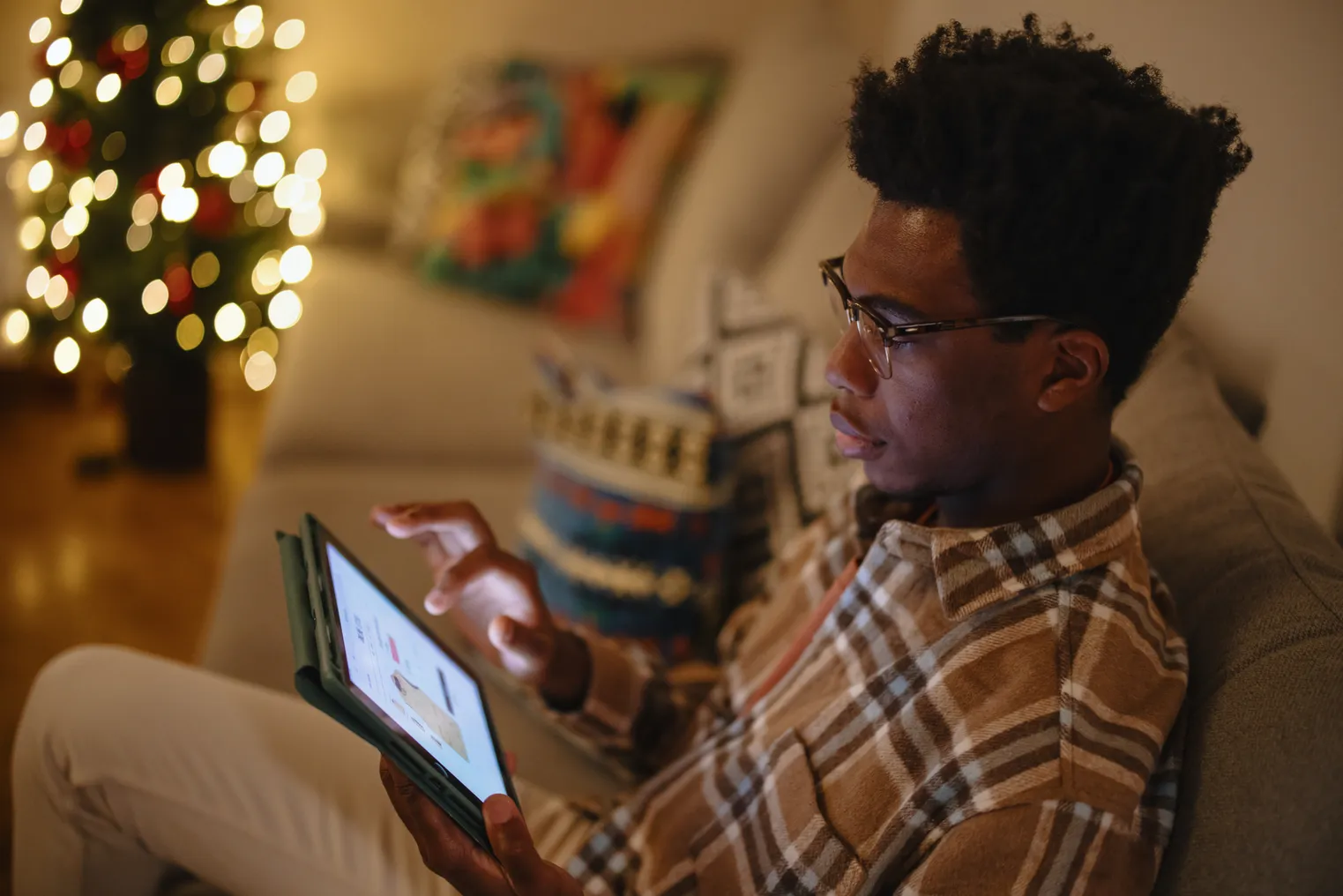 Person sits on a couch shopping on an iPad next to a Christmas tree.