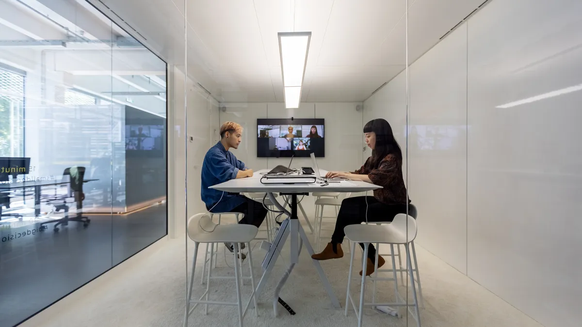 Two businesspeople sitting inside a small cubicle and working on their laptops. Man and woman working in hybrid office cubicle.