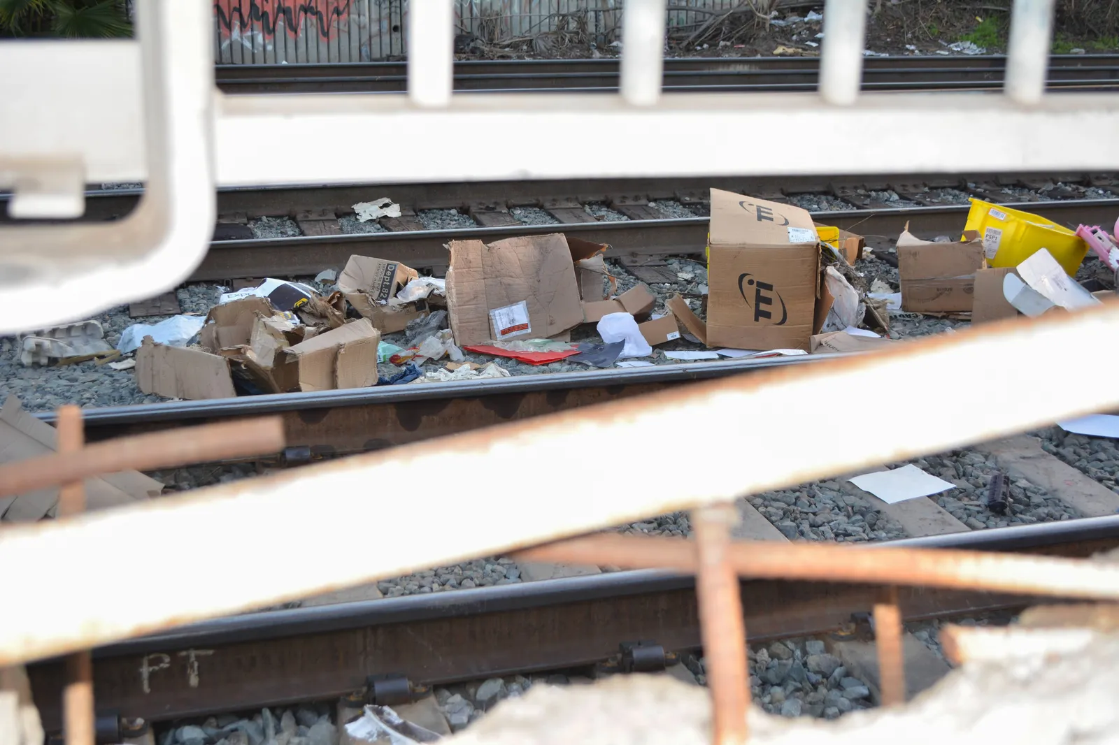 Open packages and debris on railroad tracks is seen through metal fencing.