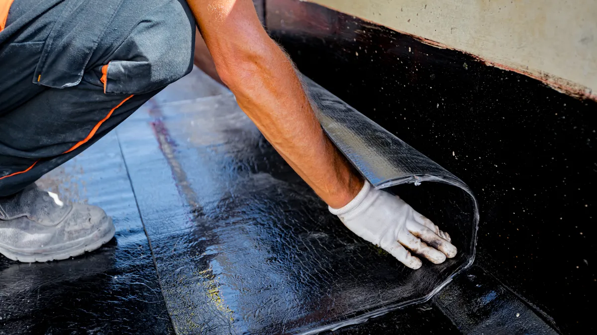 Worker unrolls foil with bitumen waterproofing installation over concrete