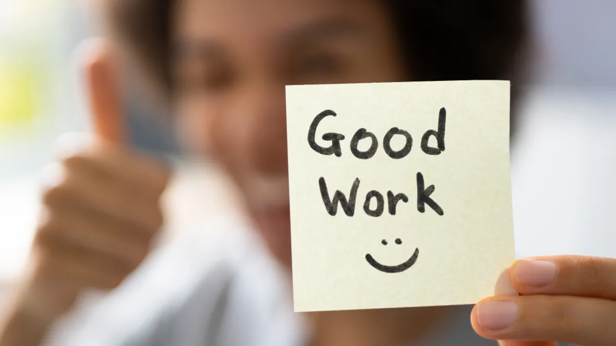 A person holding up a post it reading "Good Work" and giving a thumbs up.