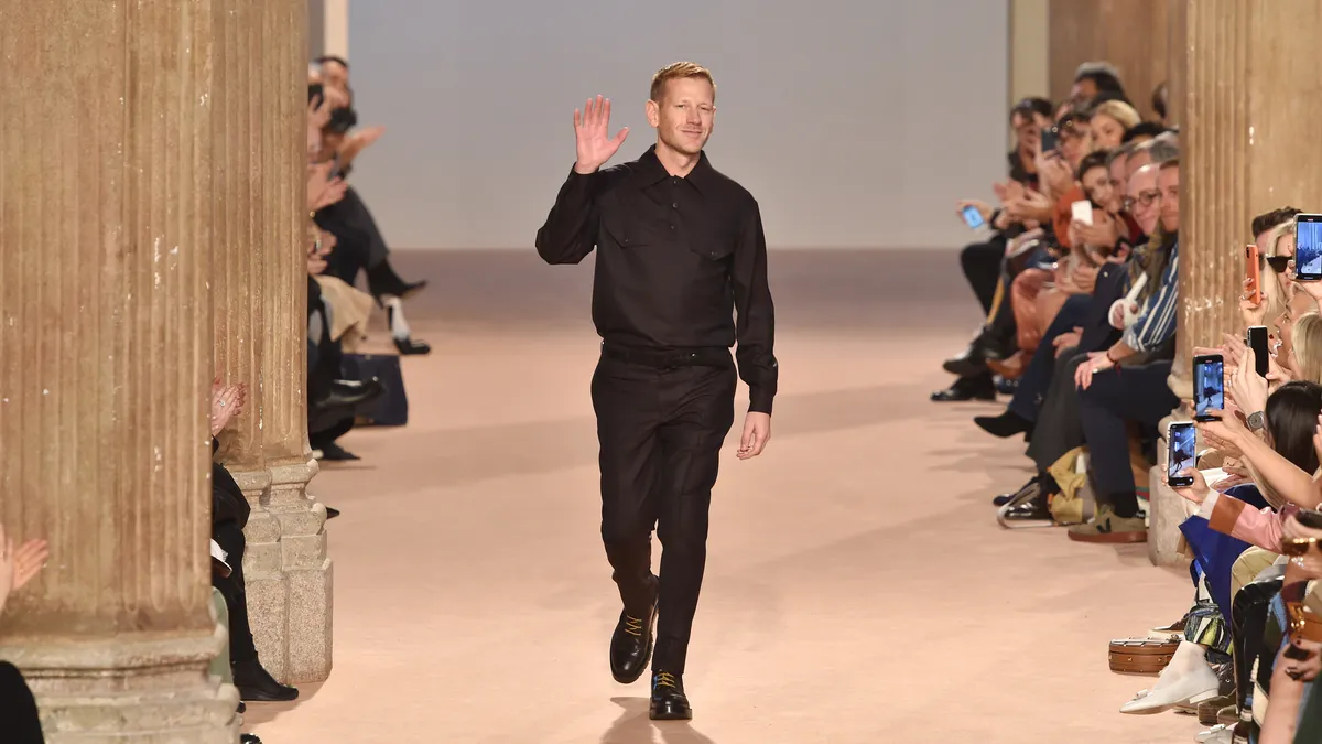 A person in black clothing waves to the audience while walking down a runway.