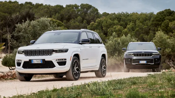 Two 2023 Jeep Grand Cherokees, one white and one black, are seen driving on a dirt road before a forested background.