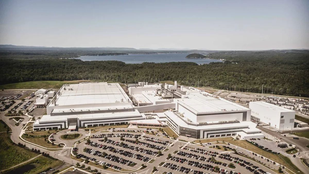 A white building with a parking lot with cars on front of it and trees and a lake behind it.