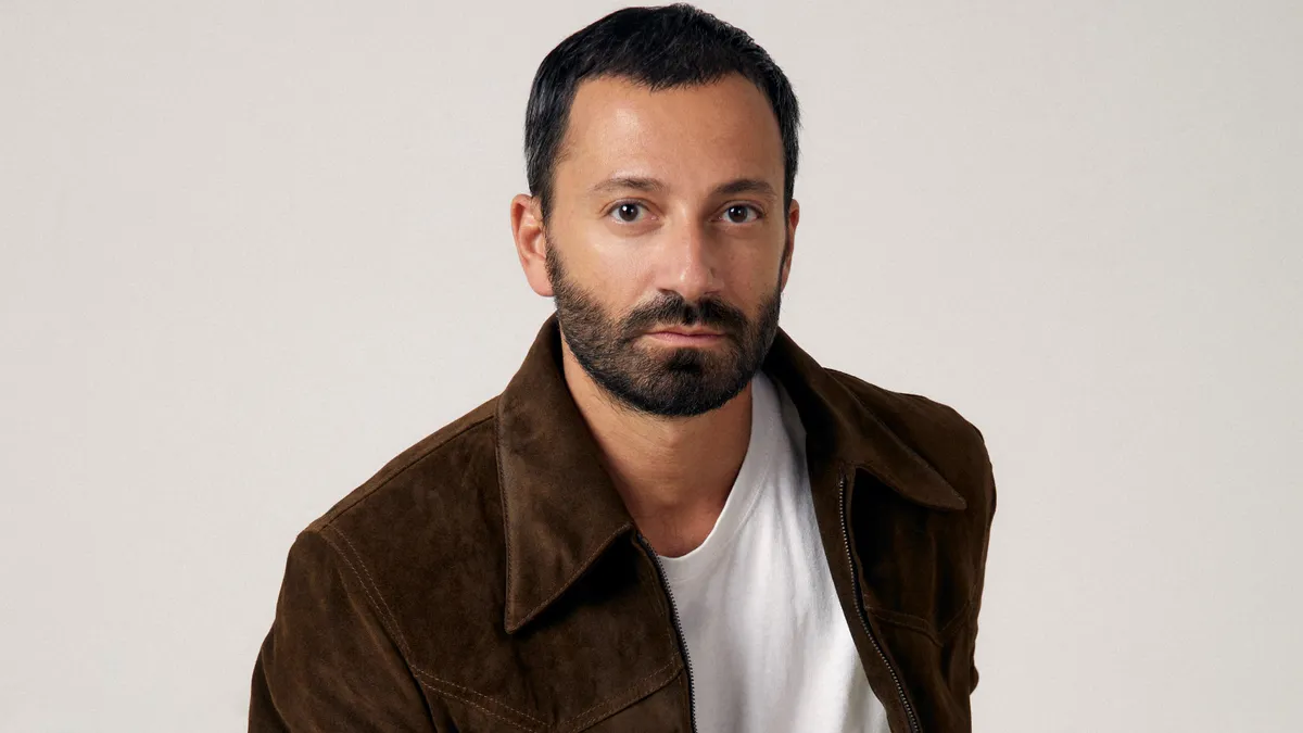 A person wearing a brown suede jacket sits and stares into the camera.