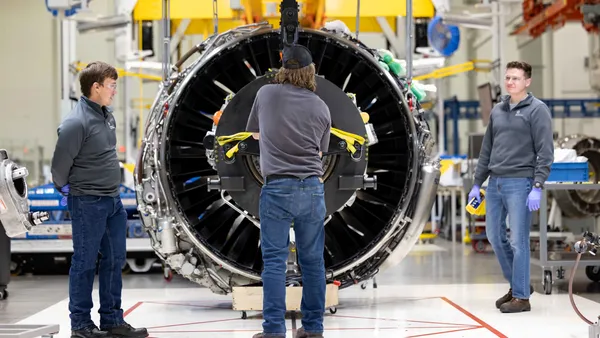 Three people in gray sweaters, blue jeans and protect eyewear working on a plane engine.