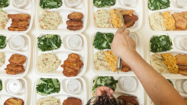 A view of a person's hand assembling multiple rows portioning meals to be served out on individual serving trays.