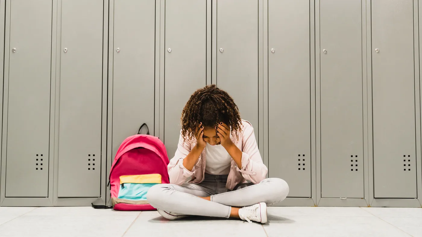 Young student sits in front of lockers inside a school building with head in hands.