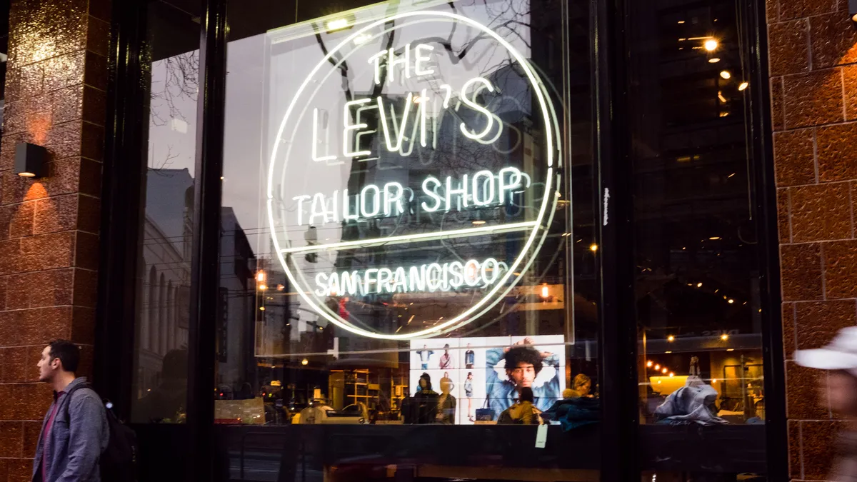 Someone stands to left of a store window with a white neon sign that reads "The Levi's Tailor Shop San Francisco."