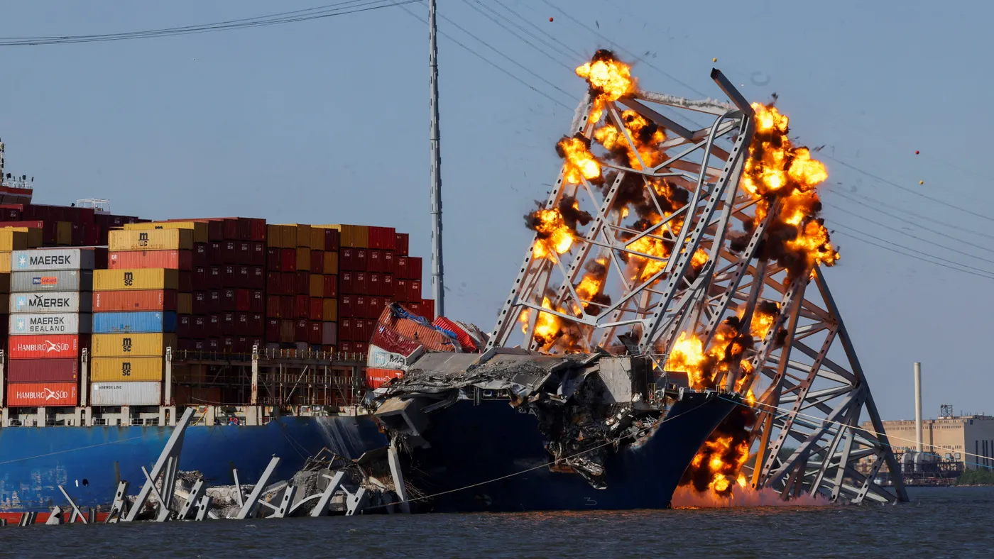 A cargo ship carrying stacked containers is trapped by a fallen bridge in flames.