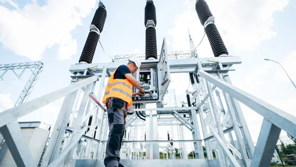 A man wearing an orange vest is positioned on a platform adjacent to a substation, monitoring the surroundings.