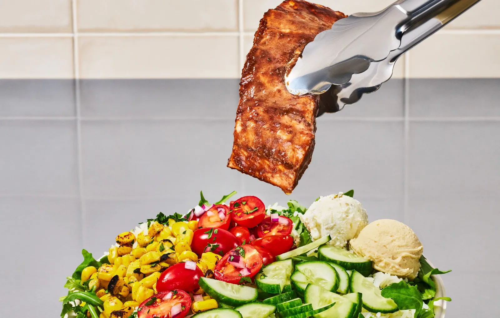 A photograph of salmon held in tongs over a bowl of various vegetables, rice and greens.