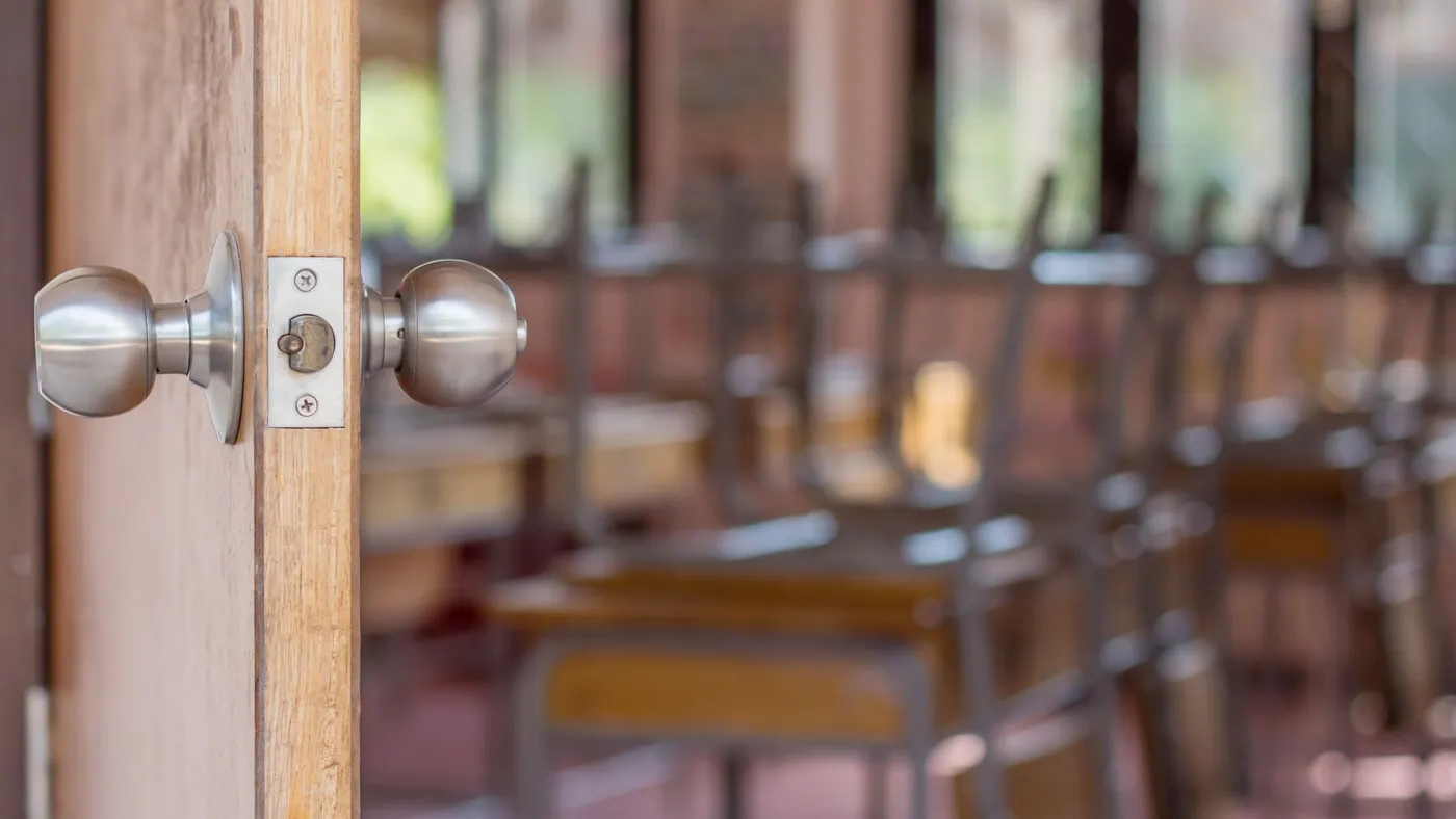 A door opens to an empty classroom with chairs flipped upside down on top of desks.