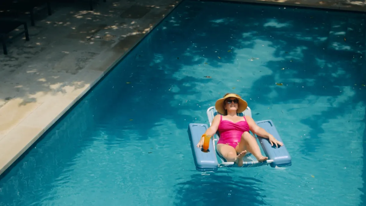 A women in a pink bathing suit and sunhat lays in a blue pool float in the middle of a pool with a cocktail.