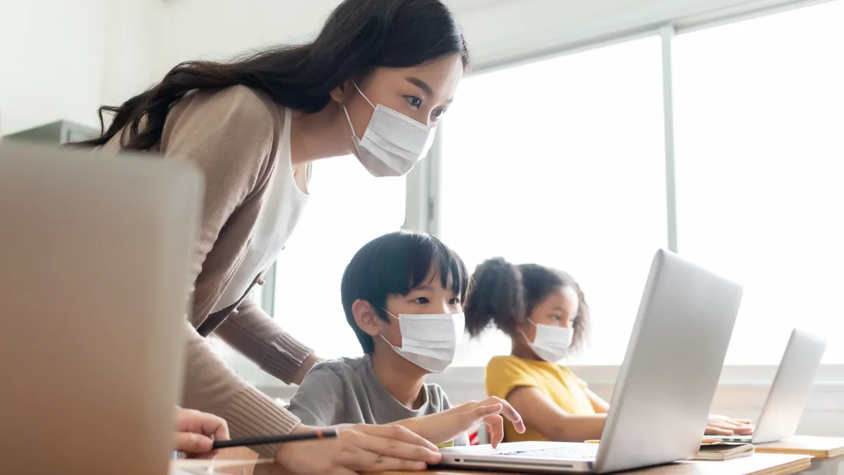 Teacher with little boy in protective face masks using laptop for studying in classroom.