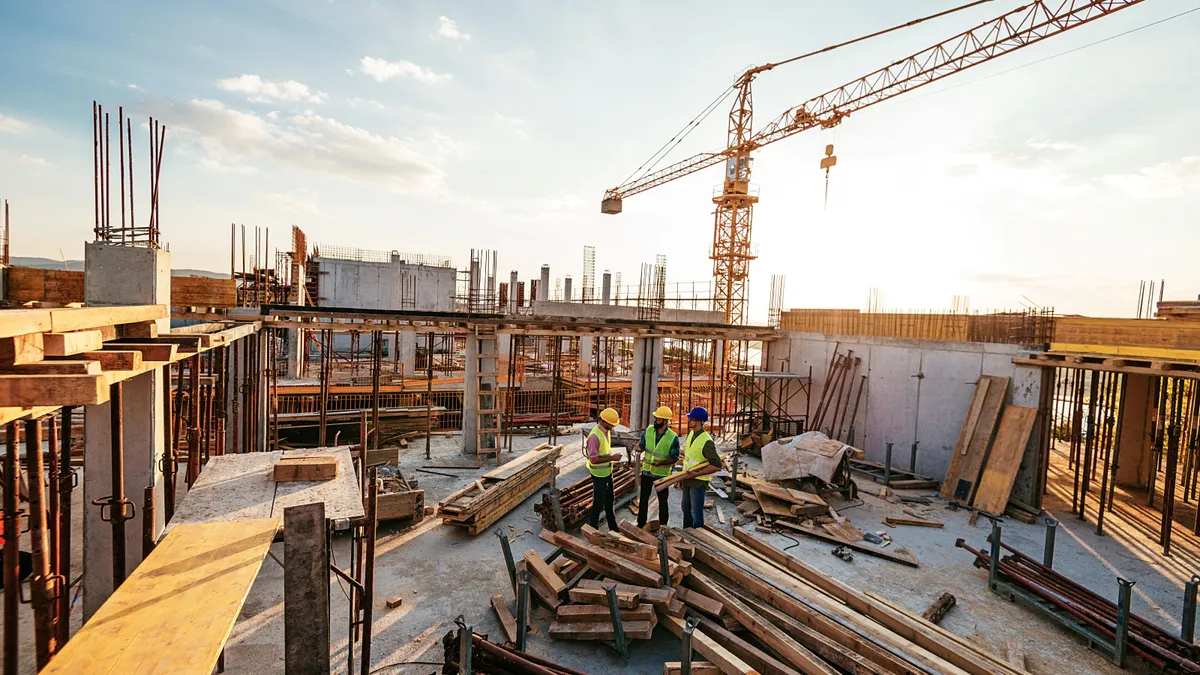 Three construction workers stand on a roof of a building under construction, surrounded by cranes and materials.