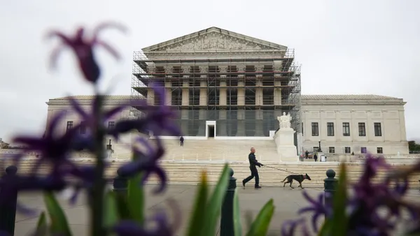 A police officer walks a dog in front of steps of columned building with scaffolding on it.