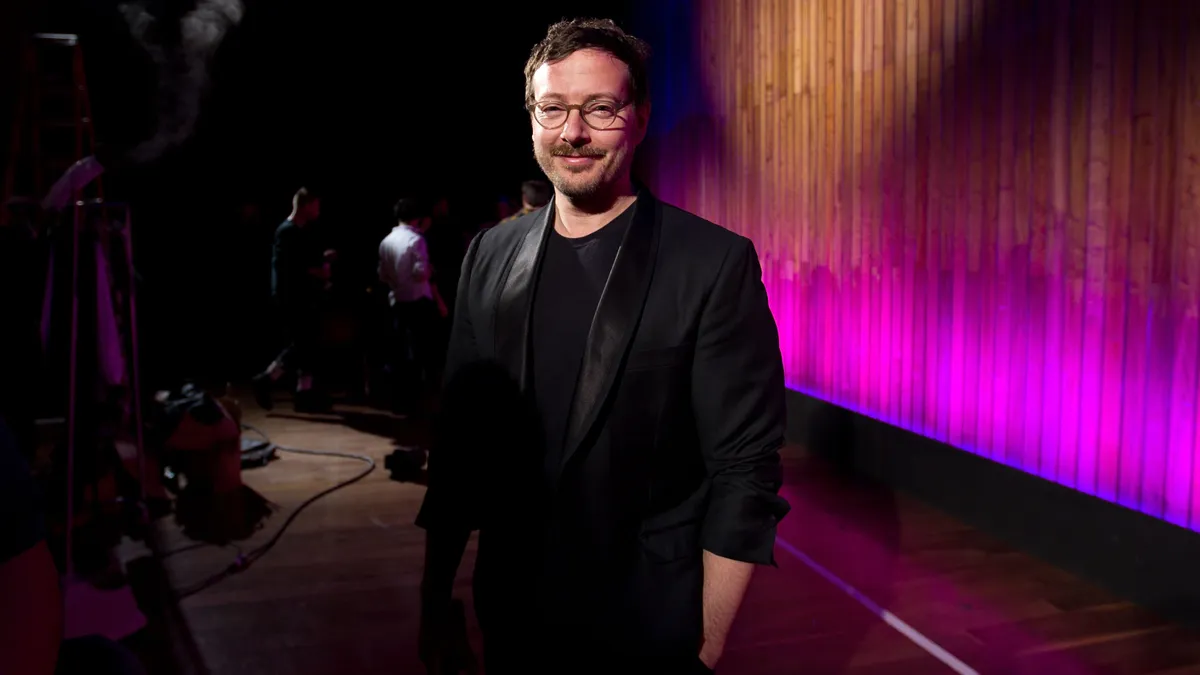 Tim Hamilton stands in front of an ombre-lit curtain wearing a black suit and black t-shirt with his hands in his pockets smiling at the camera.