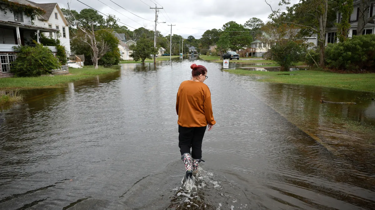 A person in tall rain boots walks in shin-deep water on a street lined by houses and power lines.