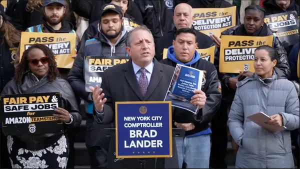 A man in a heavy coat surrounded by men and woman holding signs reading "Fair pay & safe jobs for Amazon Teamsters" and "Pass the delivery protection act."