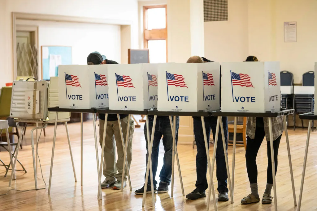 Four voting booths are lined up, filled with people.