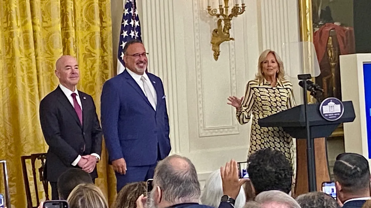 three adults stand on a stage in front of a yellow curtain. One adult is standing at a podium and speaking.