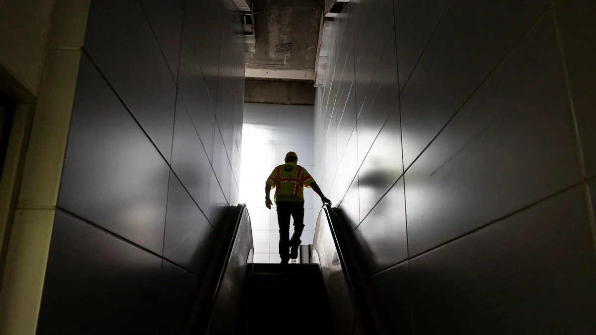 An individual, wearing a yellow safety vest, ascends an escalator.