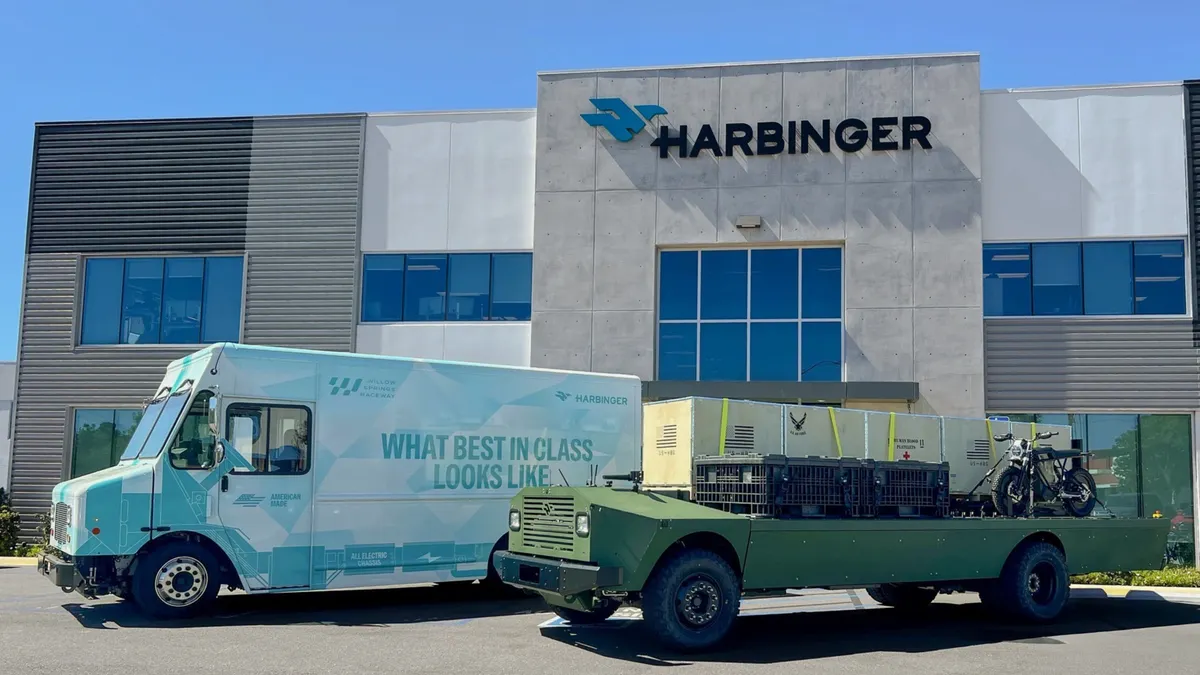 A white and light turquoise delivery truck with the Harbinger sign imprinted on it, parked next to a green military truck.