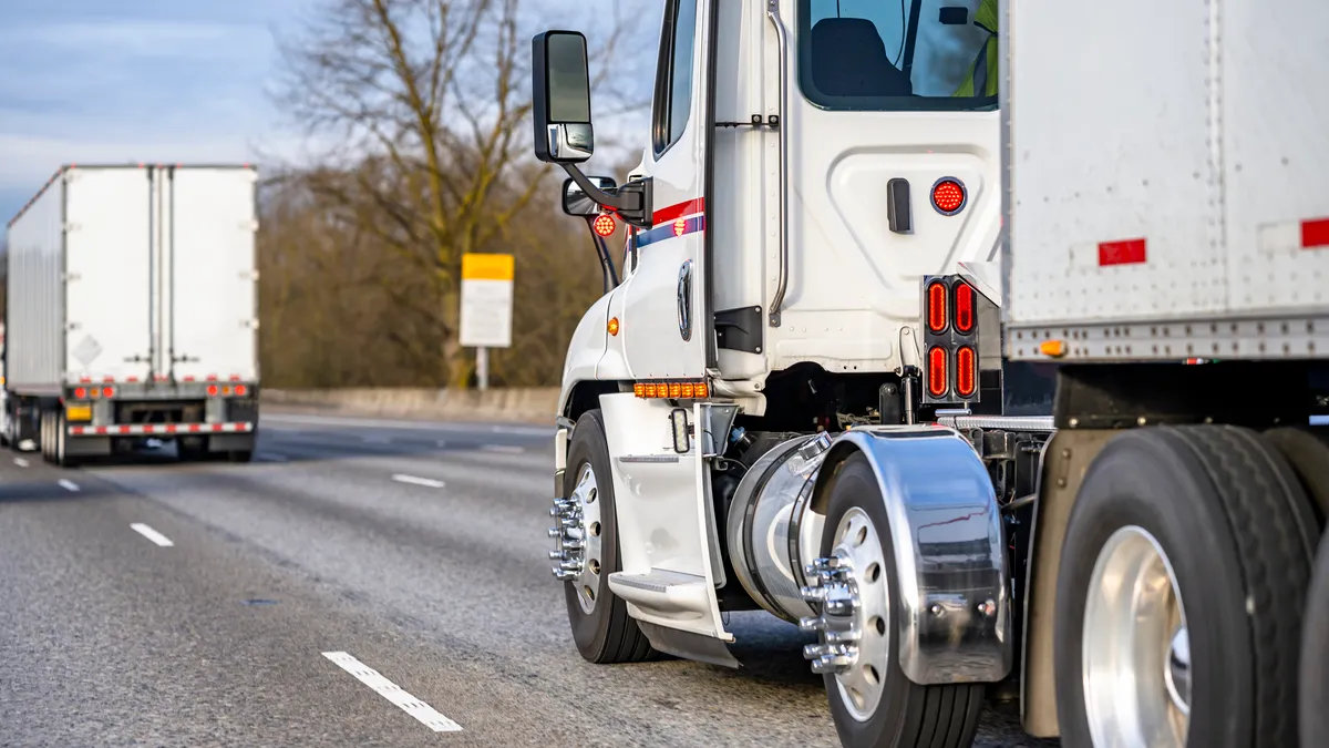 Two trucks travel on a road, as shown by a vehicle in a left lane.