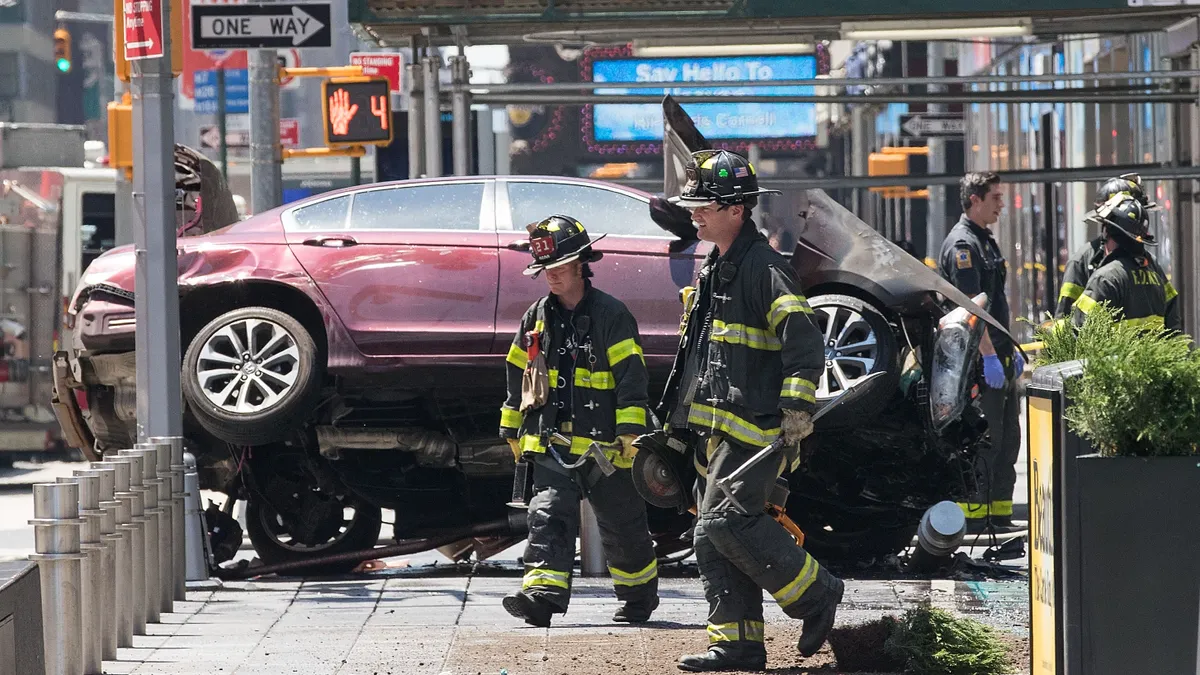 A red sedan, passenger side elevated, lying across a sidewalk in New York. A smashed, burned front end and other damage indicate it was in a crash. Firefighters are standing near it.