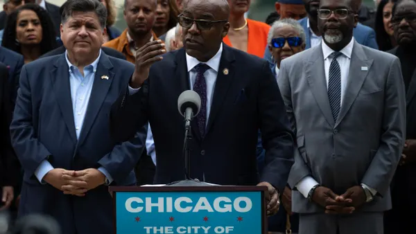 A person stands at a podium with a sign that says, "Chicago, City of Big Shoulders," surrounded by many people.