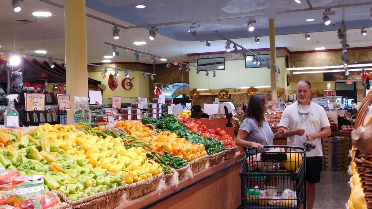 Two individuals, one man and one woman, in the produce aisle of a grocery store talking to each other