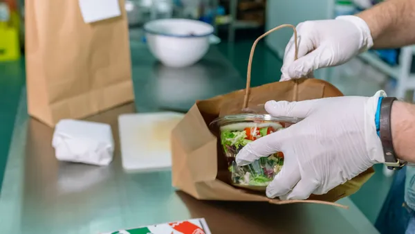 A gloved hand places a takeout salad in a paper bag