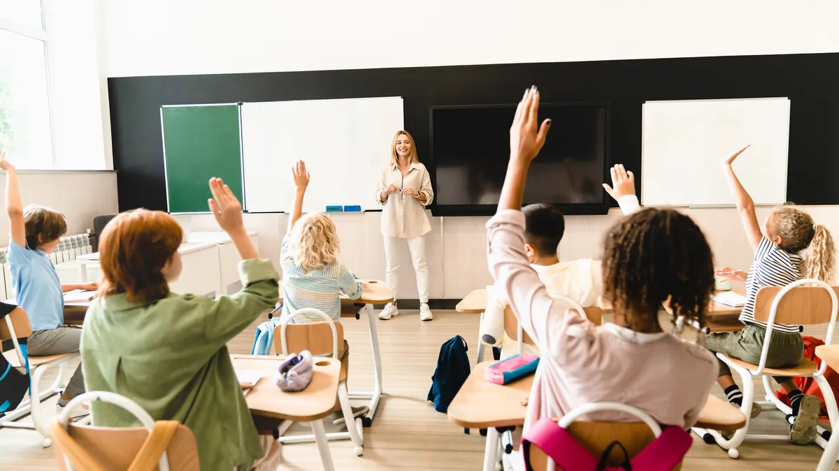 A teacher stands at the front of a classroom with a chalkboard in the background. In the foreground, students are sitting in individual desks, facing the teacher with their hands raised.