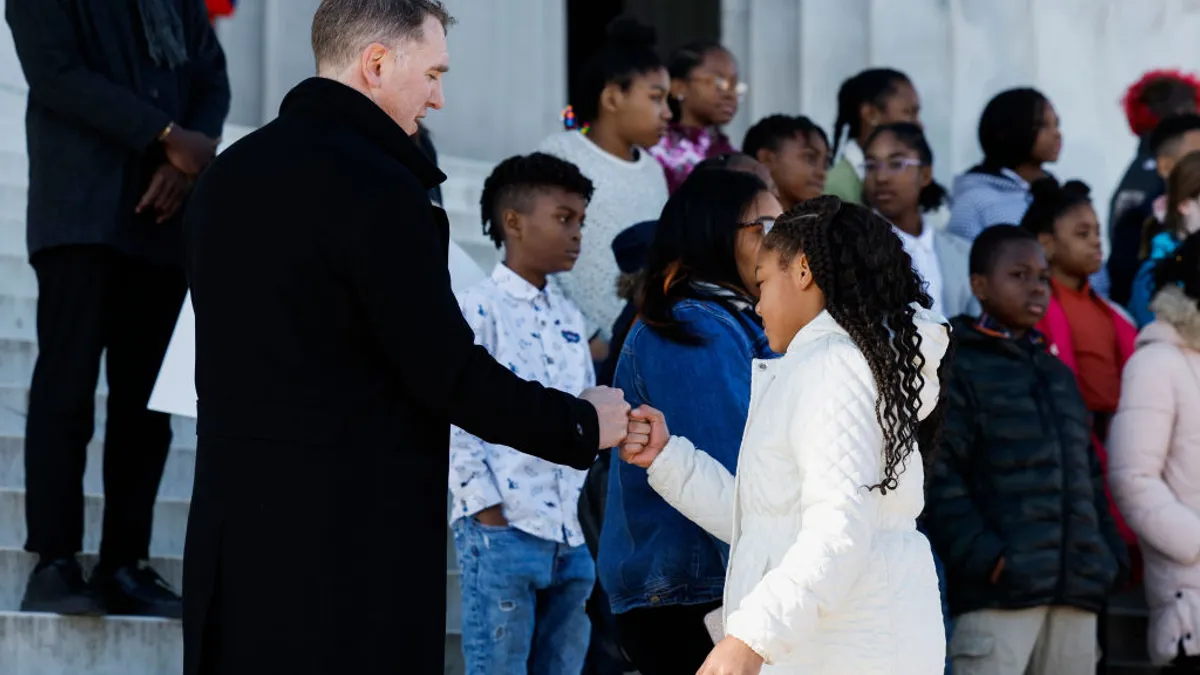 An adult and student fist bump. They are standing outside alongside other students. Everyone has jackets on.